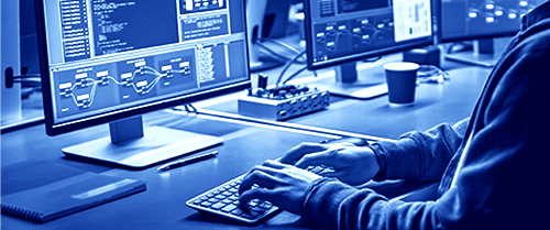 The hands of a person sitting in a high-tech lab environment at a computer terminal in front of multiple flat screens, typing.