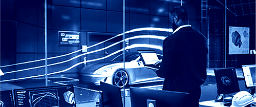 A person standing at the full-height glass wall of a modern wind tunnel laboratory, observing a vehicle being tested while the thin slipstream smoke trail flow over it.
