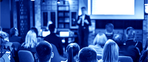 A person standing while teaching from the front of a classroom lecture hall with people in the audience, as seen from the back of the room looking forward.